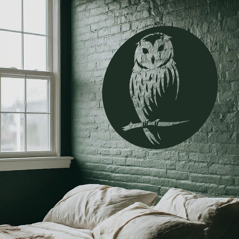 Op de afbeelding: Een zwart-witte silhouet van een uil wanddecor in een ronde vorm, gemonteerd op een groene bakstenen muur. De uil zit op een tak met zijn vleugels uitgespreid. De decoratie is boven een wit bed met twee kussens.