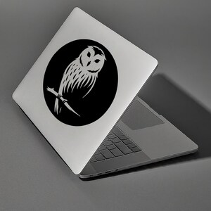 Op de afbeelding: Een zwarte en witte sticker met een silhouet van een uil op een zilveren laptop. De uil zit op een tak met uitgespreide vleugels.