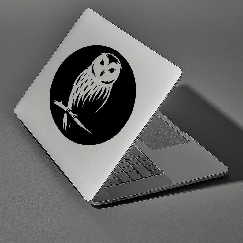 Op de afbeelding: Een zwarte en witte sticker met een silhouet van een uil op een zilveren laptop. De uil zit op een tak met uitgespreide vleugels.