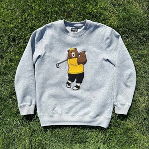 May include: A gray sweatshirt with a brown bear wearing a yellow shirt and a hat, swinging a golf club. The bear is embroidered on the front of the sweatshirt.