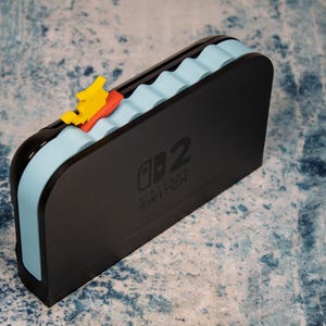Peut inclure: Station d'accueil Nintendo Switch noire avec un motif de vagues bleu clair et un bateau jaune et rouge. La station d'accueil porte le logo Nintendo Switch et le chiffre 2.
