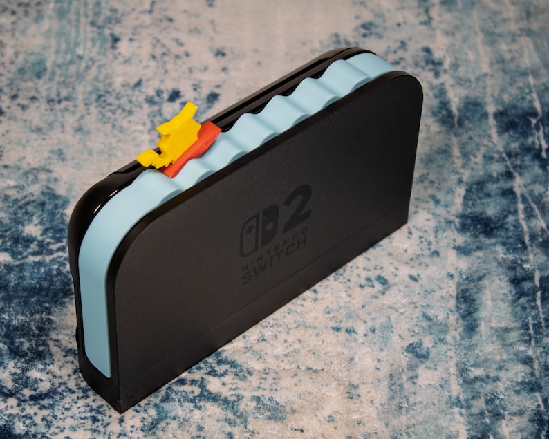 Peut inclure: Station d'accueil Nintendo Switch noire avec un motif de vagues bleu clair et un bateau jaune et rouge. La station d'accueil porte le logo Nintendo Switch et le chiffre 2.
