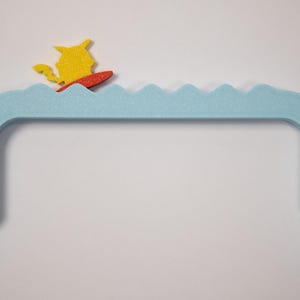 Peut inclure: Une pi&egrave;ce d&eacute;corative bleue clair, arqu&eacute;e, avec un dessus en forme de vagues. Une figurine jaune sur une planche de surf rouge est positionn&eacute;e sur les vagues. La pi&egrave;ce a une base rectangulaire.