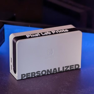 May include: A white Nintendo Switch console with a black top and the text "Pixel Lab Prints" and "PERSONALIZED" printed on it.