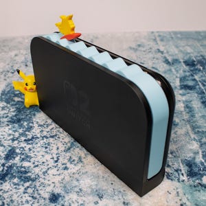 Peut inclure: Station d'accueil noire pour Nintendo Switch avec des accents bleu clair et une figurine Pikachu jaune. Le logo Nintendo Switch est visible. Une autre figurine Pikachu est visible sur le c&ocirc;t&eacute; gauche.
