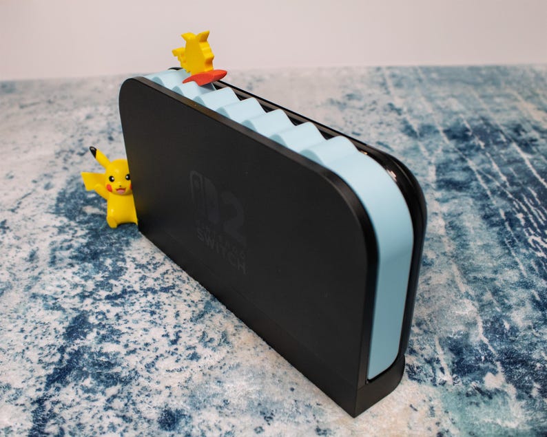Peut inclure: Station d'accueil noire pour Nintendo Switch avec des accents bleu clair et une figurine Pikachu jaune. Le logo Nintendo Switch est visible. Une autre figurine Pikachu est visible sur le c&ocirc;t&eacute; gauche.