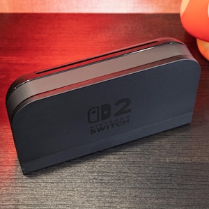 Pode incluir: Uma base preta para Nintendo Switch com o logotipo Nintendo Switch. A base tem uma ranhura para o console e um design elegante e moderno. Uma figura de cogumelo vermelho e branco e uma figura amarela de Pikachu estão no fundo.