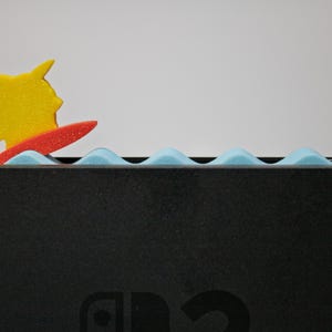 Peut inclure: Une figurine jaune de Pikachu sur une planche de surf rouge, sur fond de mousse bleue en forme de vagues. Le design est plac&eacute; sur une surface noire, peut-&ecirc;tre une console de jeu.