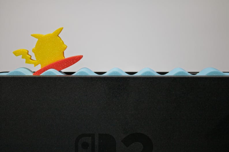 Peut inclure: Une figurine jaune de Pikachu sur une planche de surf rouge, sur fond de mousse bleue en forme de vagues. Le design est plac&eacute; sur une surface noire, peut-&ecirc;tre une console de jeu.