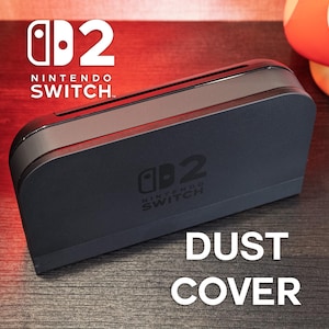 Puede incluir: Una cubierta antipolvo negra para Nintendo Switch 2 con el logotipo de Nintendo Switch. El texto "DUST COVER" se muestra en blanco. Se ve un fondo rojo y negro.