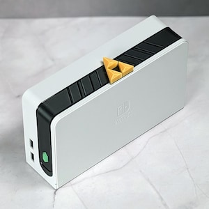 Könnte beinhalten: Ein weiß-schwarzes Nintendo Switch Dock mit einem goldenen Triforce-Symbol auf der Oberseite. Das Dock hat eine grüne Betriebsanzeige und zwei USB-Anschlüsse.