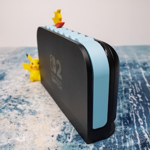 Peut inclure: Station d'accueil Nintendo Switch noire avec des accents bleu clair et le texte "Nintendo Switch". Une figurine Pikachu jaune est sur le c&ocirc;t&eacute;, et une figurine jaune et rouge est sur le dessus. La station d'accueil est sur une surface bleue et blanche.