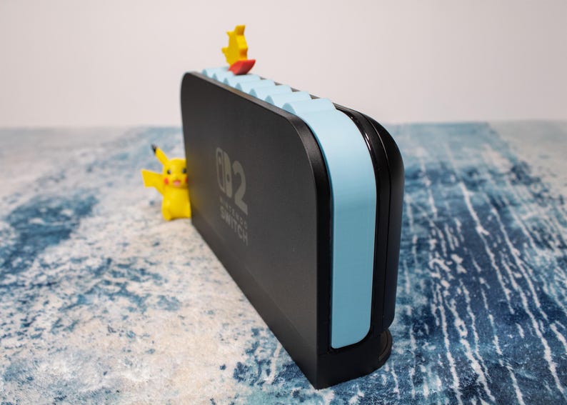 Peut inclure: Station d'accueil Nintendo Switch noire avec des accents bleu clair et le texte "Nintendo Switch". Une figurine Pikachu jaune est sur le c&ocirc;t&eacute;, et une figurine jaune et rouge est sur le dessus. La station d'accueil est sur une surface bleue et blanche.