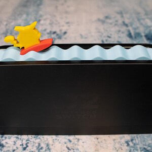 Peut inclure: Station d'accueil Nintendo Switch noire avec un dessus bleu clair en forme de vagues. Une figurine de dessin anim&eacute; jaune et rouge sur une planche de surf est fix&eacute;e au sommet. Le logo Nintendo Switch est visible &agrave; l'avant.