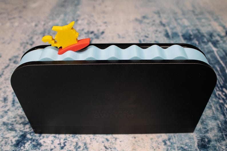 Peut inclure: Station d'accueil Nintendo Switch noire avec un dessus bleu clair en forme de vagues. Une figurine de dessin anim&eacute; jaune et rouge sur une planche de surf est fix&eacute;e au sommet. Le logo Nintendo Switch est visible &agrave; l'avant.
