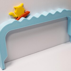 Peut inclure: Une poign&eacute;e en mousse bleu clair avec un motif de vagues, repr&eacute;sentant un surfeur jaune et orange. La poign&eacute;e a une forme incurv&eacute;e et est con&ccedil;ue pour &ecirc;tre fix&eacute;e au mur. Le surfeur est sur une petite planche de surf orange.