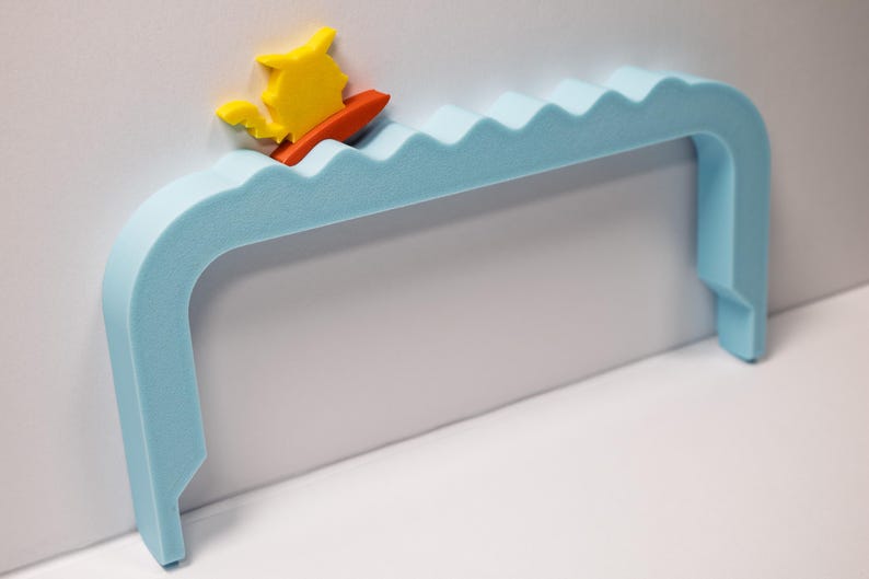 Peut inclure: Une poign&eacute;e en mousse bleu clair avec un motif de vagues, repr&eacute;sentant un surfeur jaune et orange. La poign&eacute;e a une forme incurv&eacute;e et est con&ccedil;ue pour &ecirc;tre fix&eacute;e au mur. Le surfeur est sur une petite planche de surf orange.