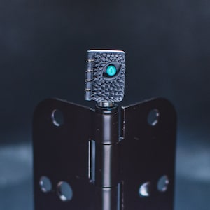 May include: A black door hinge with a small, black, textured book charm with a turquoise eye on top.
