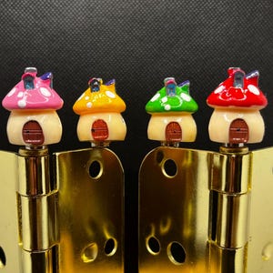Pode incluir: Quatro puxadores decorativos em forma de cogumelo em rosa, amarelo, verde e vermelho com bolinhas brancas. Os puxadores são montados em dobradiças douradas, criando um toque lúdico e colorido.