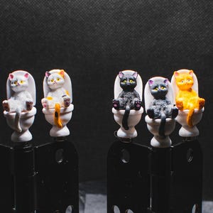 Peut inclure: Cinq figurines de chat en céramique assises sur des toilettes. Les chats sont dans différentes poses et couleurs, notamment blanc, noir, orange et calico.
