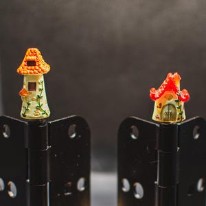 May include: Two miniature fairy houses, one with an orange roof and the other with a red roof, are attached to black metal hinges. The houses are decorated with white paint and have small windows and doors.