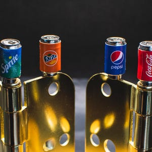 May include: Four miniature soda cans attached to gold door hinges. The cans are labelled "Sprite", "Fanta", "Pepsi", and "Coca-Cola".