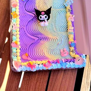 Puede incluir: Una funda para tableta decorada con patrones texturizados y ondulados en tonos pastel morado, azul y amarillo. El borde presenta remolinos de glaseado amarillo y azul adornados con pequeños dijes de animales de plástico de colores y estrellas, con una figura central de Kuromi.