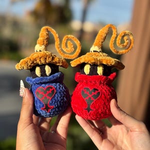 May include: Two plush figures, one blue and one red, each with a black heart design. They have black faces, yellow hats with curly accents. The figures are held up against a blurred background.