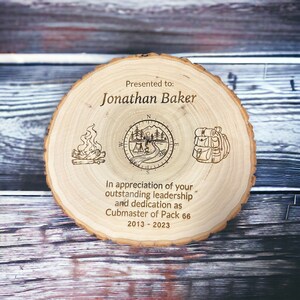Puede incluir: Una placa de madera con un dise&ntilde;o de br&uacute;jula, fogata y mochila. Dice "Presentado a: Jonathan Baker" y "En reconocimiento a su liderazgo sobresaliente y dedicaci&oacute;n como Cubmaster de la Manada 66 2013-2023".