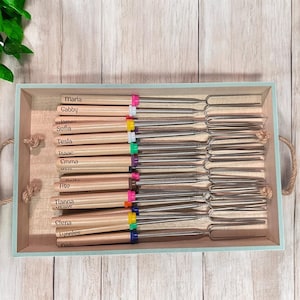 Puede incluir: Una caja de madera llena de 15 palos de asar malvaviscos de madera personalizados con puntas de colores. Los palos están dispuestos en filas y etiquetados con nombres como Maria, Gabby, Sofia, Tesla, Isaac, Emma, y más.