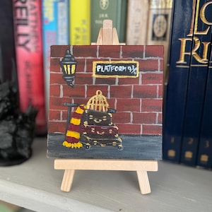 May include: A square canvas painting on a wooden easel depicting a red brick wall with a sign reading PLATFORM 9 3/4. The scene includes a black wall lamp, a stack of dark suitcases, a gold birdcage, and a red and yellow striped scarf.