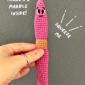 May include: A pink and orange crocheted worm toy with a marble inside. The toy features a cartoon face with black and white eyes and a small mouth. Text on the image reads: THERE'S A MARBLE INSIDE! and SQUEEZE ME. A logo for fox creations is visible.