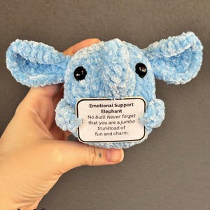 Peut inclure: Un éléphant en peluche bleu clair crocheté avec des yeux noirs. Le jouet tient une carte blanche avec le texte "Emotional Support Elephant No bull! Never forget that you are a jumbo trunkload of fun and charm."