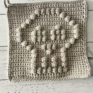 May include: A beige crochet pot holder with a textured design. The pot holder has a loop for hanging.