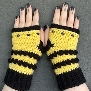 May include: A pair of yellow and black crochet fingerless gloves. The gloves feature a bumblebee design with black stripes and a black cuff. The top edge is trimmed with black and yellow beads. The gloves are handmade.