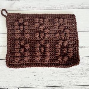 May include: A rectangular dark brown crochet potholder featuring the raised bobble stitch text "HOT STUFF" centered on the front. The item includes a small crocheted loop at the top left corner for hanging, crafted with a consistent yarn texture.