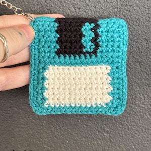 May include: Crocheted teal floppy disk keychain with black and white details. The keychain has a silver chain and the text "fox creations" in the bottom right corner.