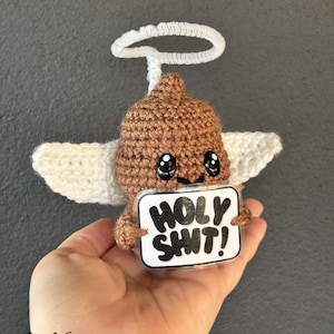 Puede incluir: Un emoji de caca marrón tejido a ganchillo con alas blancas y un halo. El emoji sostiene un letrero que dice "HOLY SHIT!" en texto negro. El artículo está hecho a mano y tiene un diseño caprichoso.