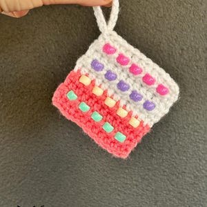 May include: A square crochet ornament featuring a white top section with pink and purple beads, and a coral bottom section with cream and mint green beads. The piece includes a white yarn loop at the top for hanging, with a fox creations logo below.