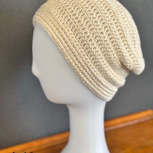 May include: A cream-colored, crocheted beanie hat. The hat is displayed on a white mannequin head. The hat has a ribbed brim and a loose, slouchy fit. The hat is made of a soft, textured yarn. The logo "fox creations" is visible in the bottom left corner.