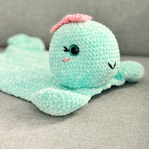 Può includere: Una coperta e un giocattolo per bambini fatti a mano a forma di balena. La balena è azzurra con una pinna rosa e tratti del viso ricamati. La coperta è morbida e strutturata.