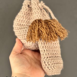 May include: A small crocheted drawstring bag with a brown tassel. The bag is light pink and has a simple, textured design.