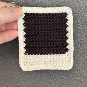 May include: A crocheted polaroid-style keychain. The central area is black, surrounded by a white border. A silver metal ring is attached for hanging. The text "fox creations" is displayed in the bottom left corner of the image.