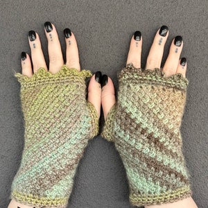 May include: A pair of handmade crochet fingerless gloves. The gloves feature a mix of green, brown, and tan yarn in a diagonal pattern. The cuffs have a scalloped edge. The Fox Creations logo is visible in the bottom left corner.