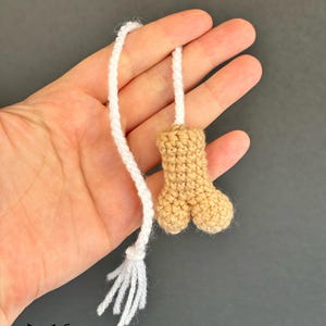 May include: A crocheted dog bone toy with a white tassel. The bone is tan and the tassel is made of white yarn.