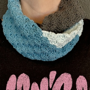 May include: A handmade crochet cowl in shades of blue, light blue, and gray. The cowl is worn around the neck and is made of a soft, textured yarn. The bottom of the image shows a black t-shirt with pink text and a fox logo.
