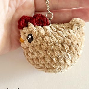 Puede incluir: Un llavero de pollo de ganchillo beige con una cresta roja de ganchillo y ojos negros. El llavero está unido a una cadena plateada.