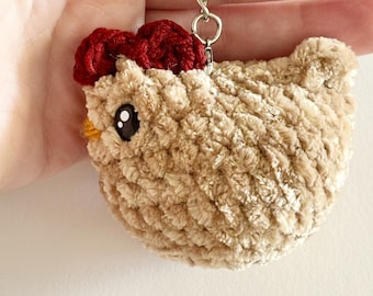 May include: A beige crocheted chicken keychain with a red crocheted comb and black eyes. The keychain is attached to a silver chain.