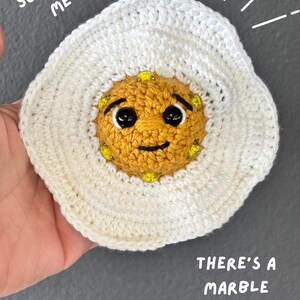 Könnte beinhalten: Ein gehäkeltes Spiegelei-Spielzeug mit weißem Rand und goldgelber Mitte, das ein lächelndes Gesicht, schwarze Kunststoffaugen und gelbe Perlen aufweist. Der Text SQUEEZE ME und THERE'S A MARBLE INSIDE! ist auf dem Bild zu sehen.