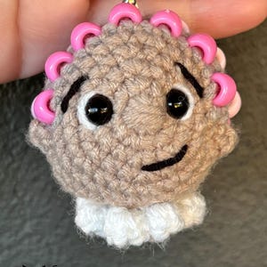 Op de afbeelding: Gebreide sleutelhanger met een bruin gezicht, roze kralen rond het hoofd en wit gebreid haar. De sleutelhanger is bevestigd aan een zilveren ketting.